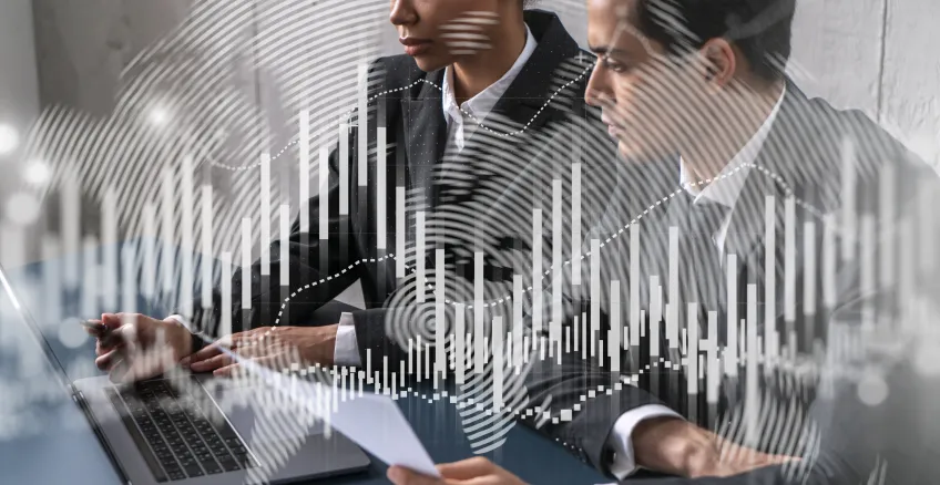 Two professionals working together at a desk, reviewing documents on a laptop, with abstract data charts and digital graphics overlaid, symbolising analysis, reporting and data-driven decision-making.